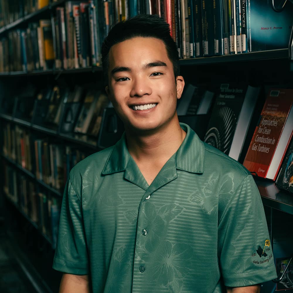 Alexander poses in front of a library bookshelf. He has medium skin tone and wears a green shirt with a subtle hibiscus pattern.