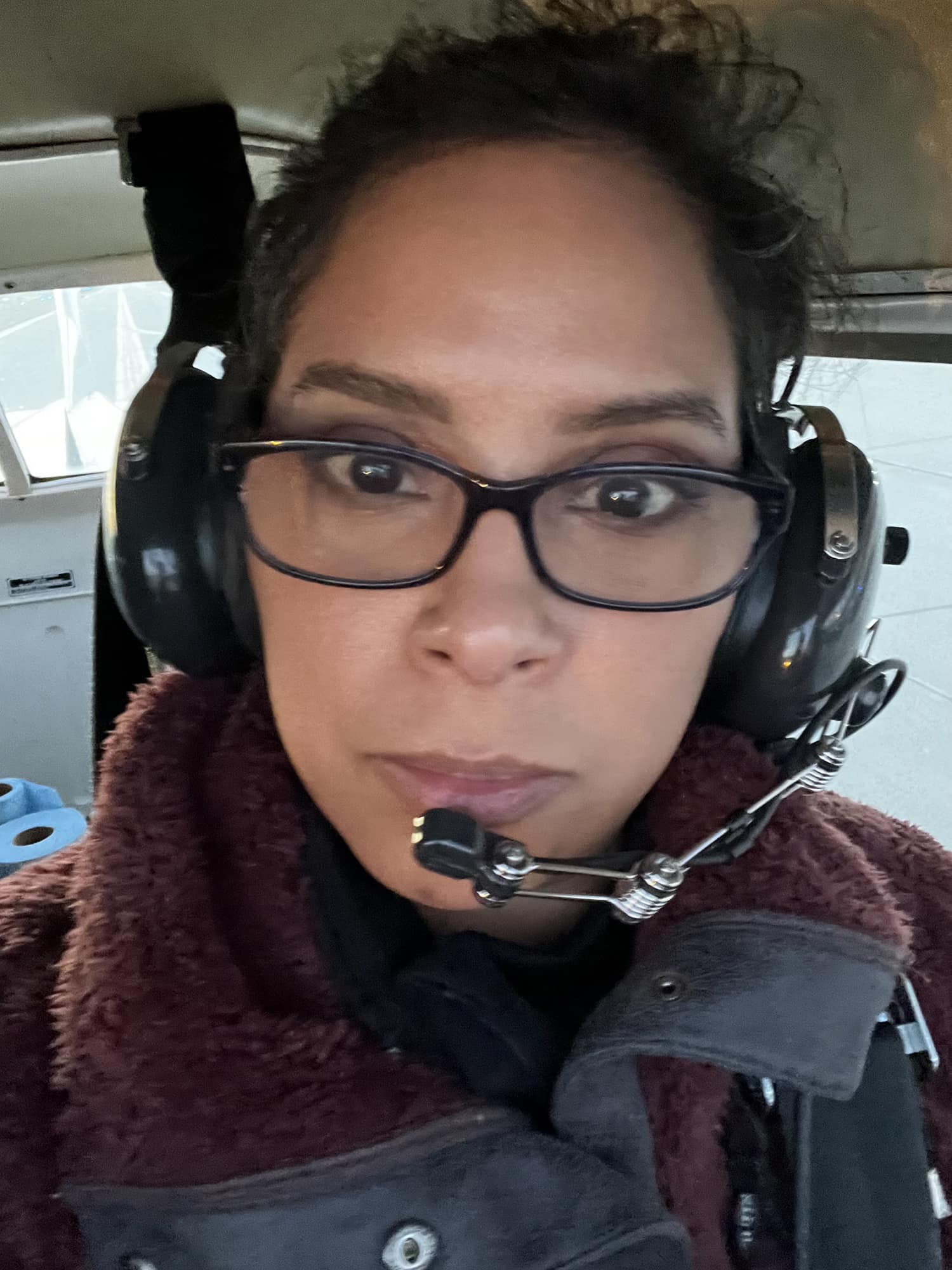 Closeup of Evelyn in the cockpit of a small plane wearing a pilot headset and a coat with a cowl collar.
