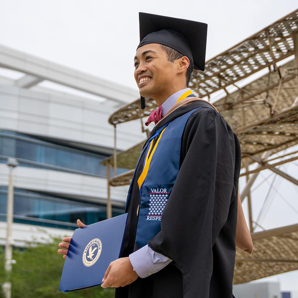 East Asian man wearing graduation cap and gown with a hood and a blue and yellow sash that reads Valor - Respect. He holds up his diploma cover for a photo in front of the historical biplane replica on the Daytona Beach Campus.