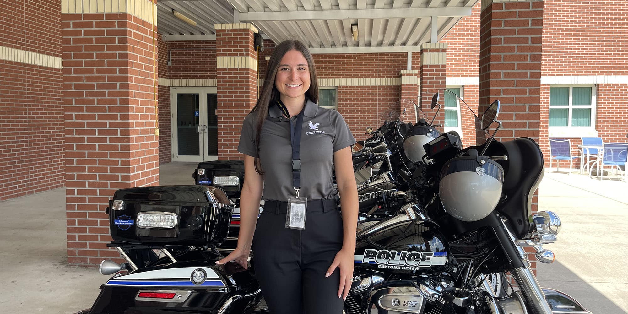 Hannah, a woman with light skin tone and long brown hair, stands in front of a line of parked motorcycles with Daytona Beach Police imprint. She's wearing a grey polo with an Embry-Riddle eagle and an id on a lanyard.