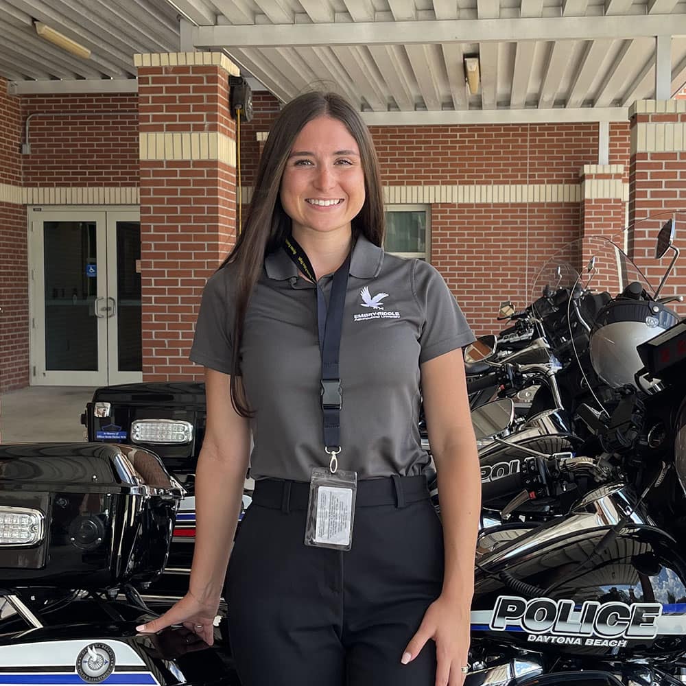 Hannah, a woman with light skin tone and long brown hair, stands in front of a line of parked motorcycles with Daytona Beach Police imprint. She's wearing a grey polo with an Embry-Riddle eagle and an id on a lanyard.