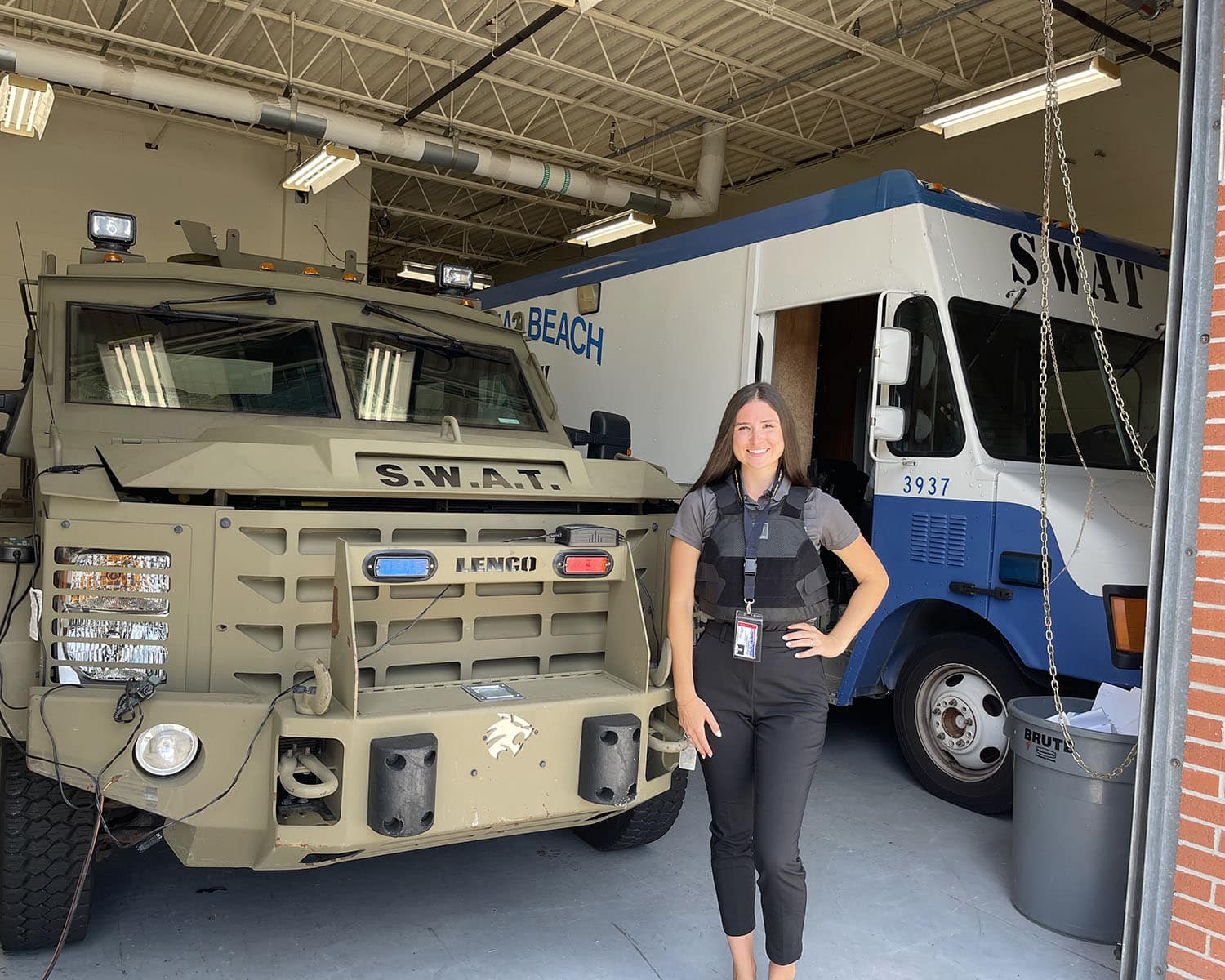 Hannah stands in a parking bay in front of two SWAT vehicles - one is a khaki-colored armored Lenco BearCat and the other is a blue and white delivery vehicle.
