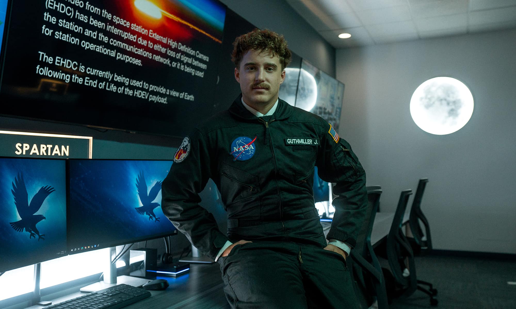A student with a mustache wearing a jumpsuit with a NASA insignia leans casually against a desk with several computer monitors in a darkened room. A glowing moon shines on the wall behind him.