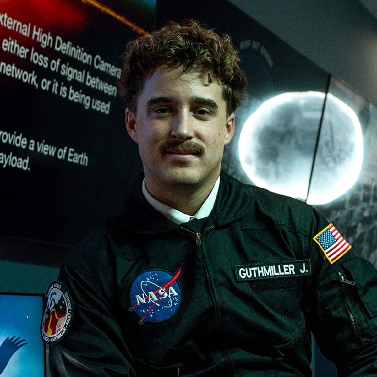 A student with a mustache wearing a jumpsuit with a NASA insignia. A glowing moon shines on the wall behind him.