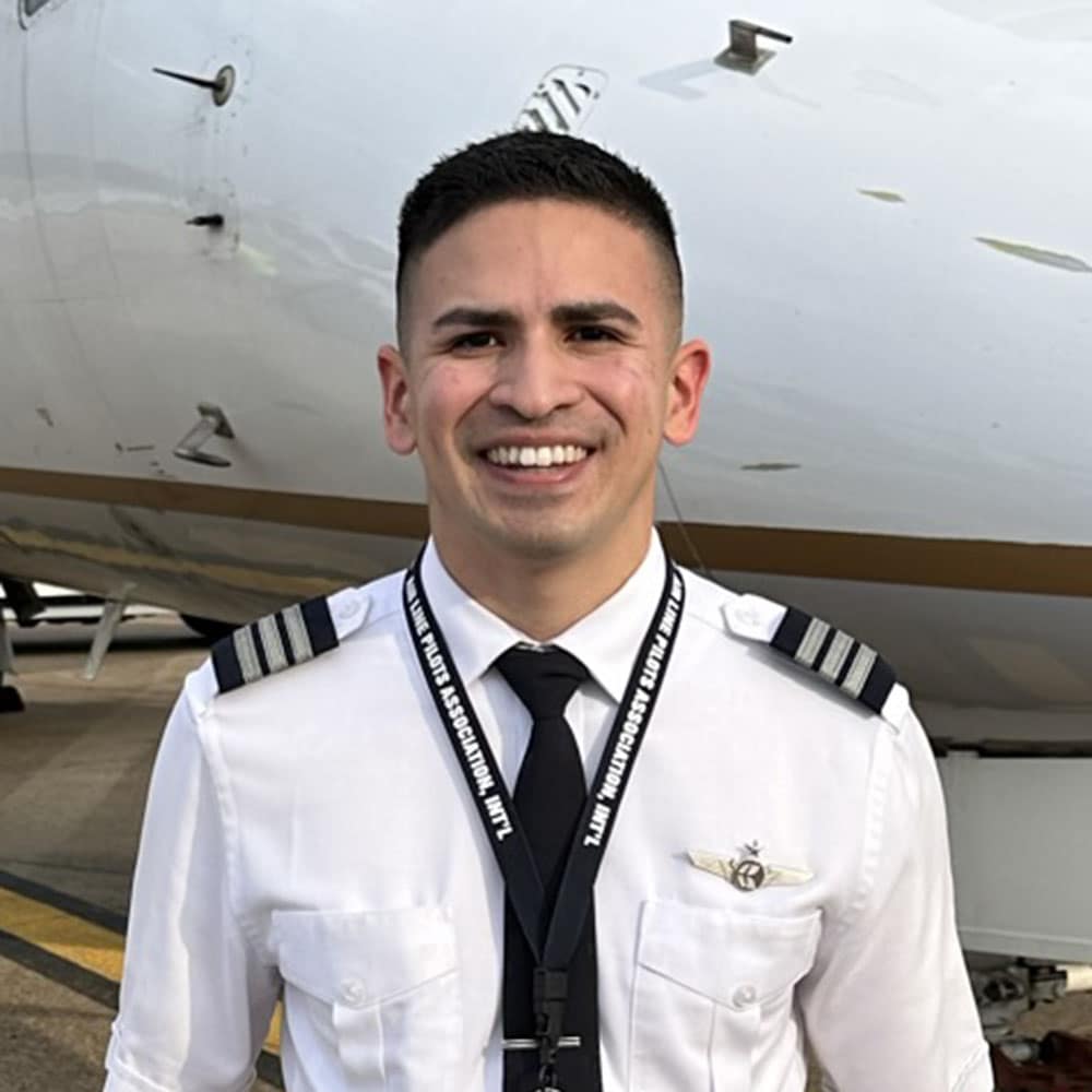 Juan, a man with medium skin tone, wears a pilot uniform and stands in front of a small corporate jet.