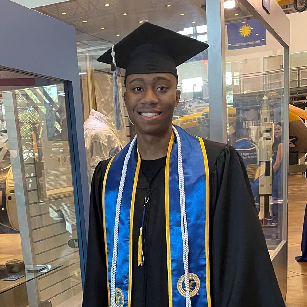 Kegan, a man with dark skin tone, wears a graduation cap and gown, a blue and yellow sash, and white cords.