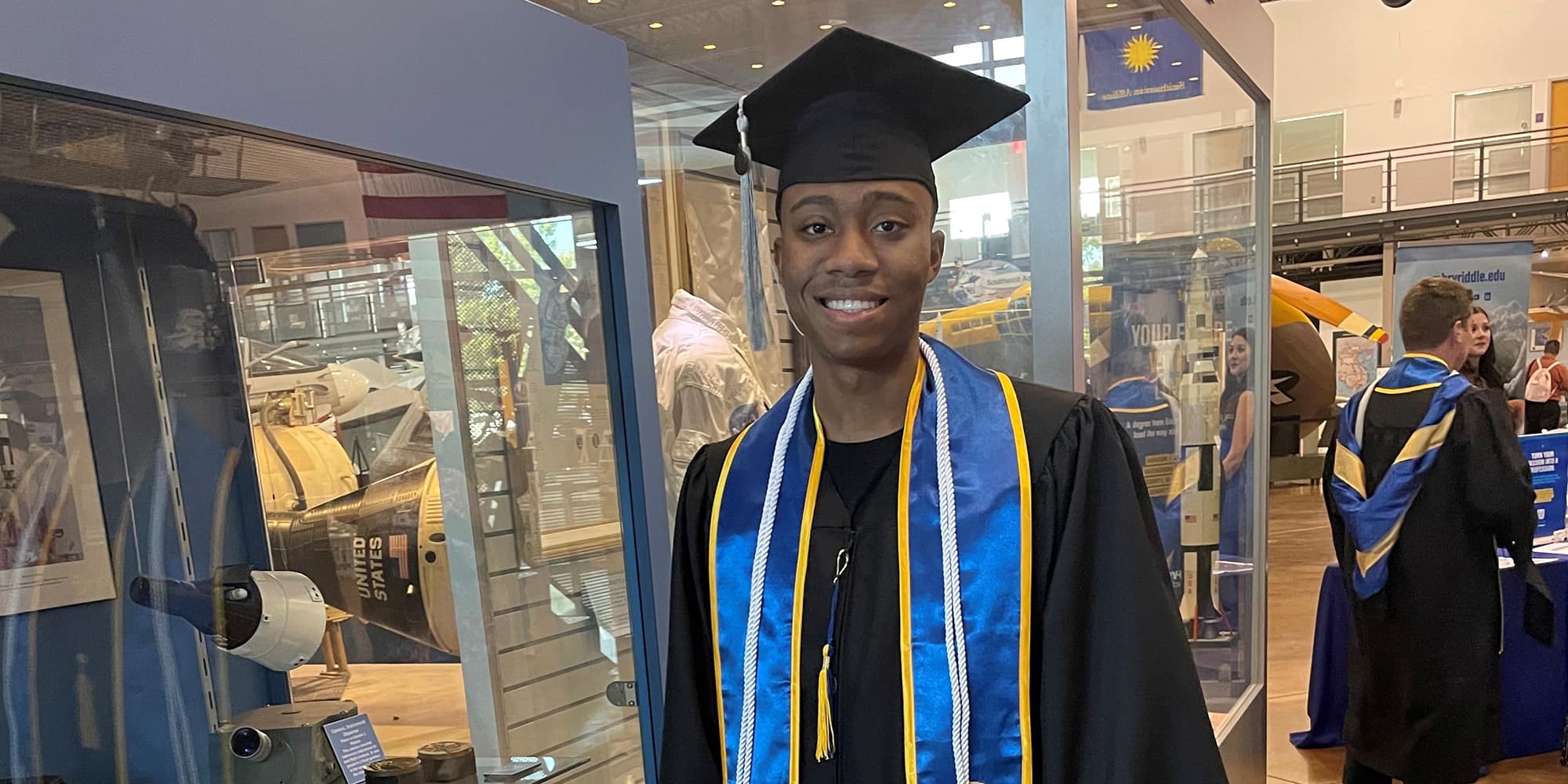 Kegan, a man with dark skin tone, wears a graduation cap and gown, a blue and yellow sash, and white cords.