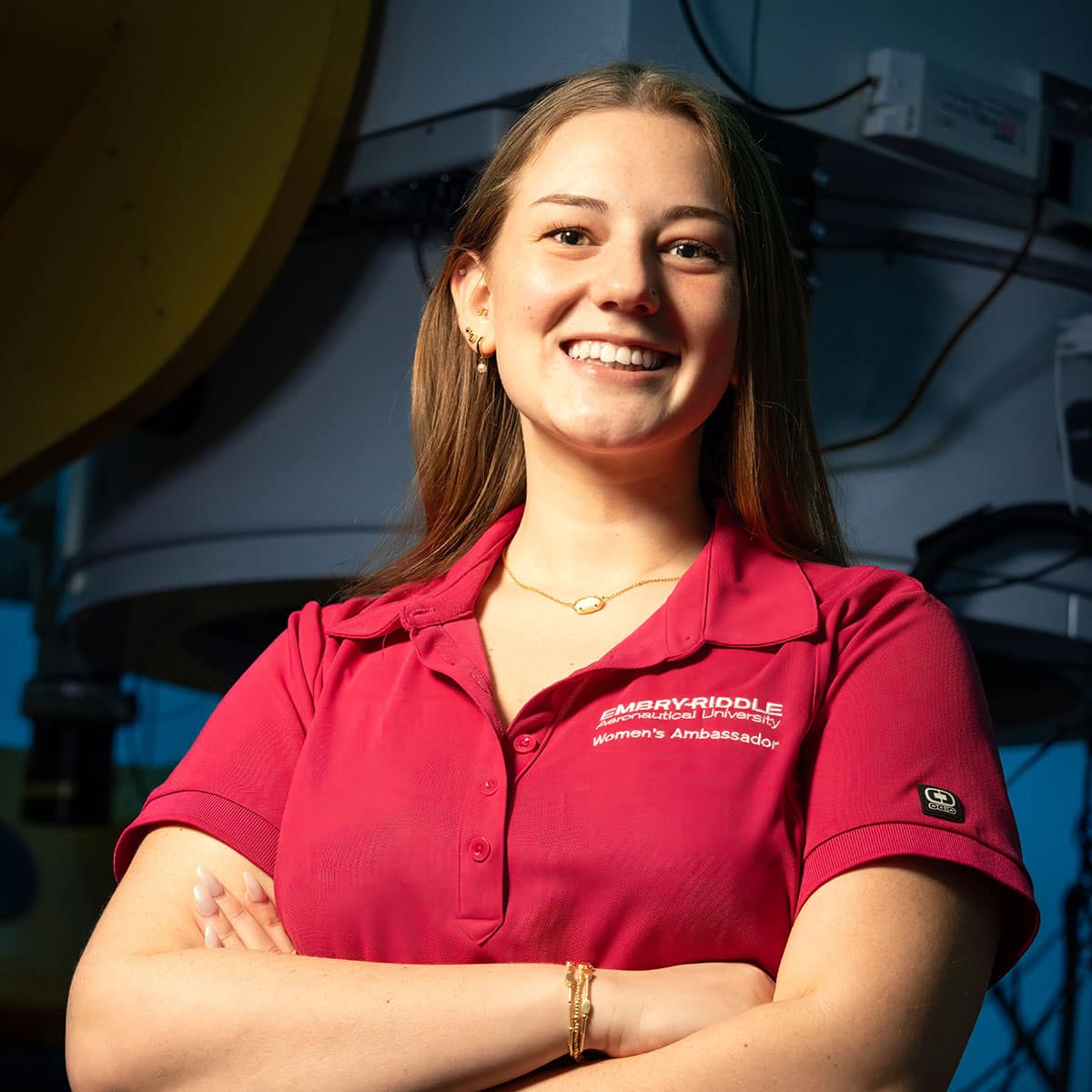 Logan, a white woman with dark blond hair pulled back, poses with her arms crossed, wearing a red polo shirt with the Embry-Riddle wordmark.