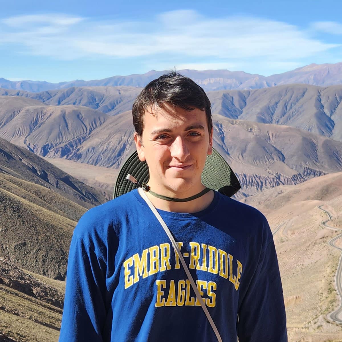 Zackrey poses, squinting into the camera, with a background of brown and gold canyon stretching into the distance. He has light skin tone and is wearing an Embry-Riddle Eagles sweatshirt and a safari hat hanging around his neck.
