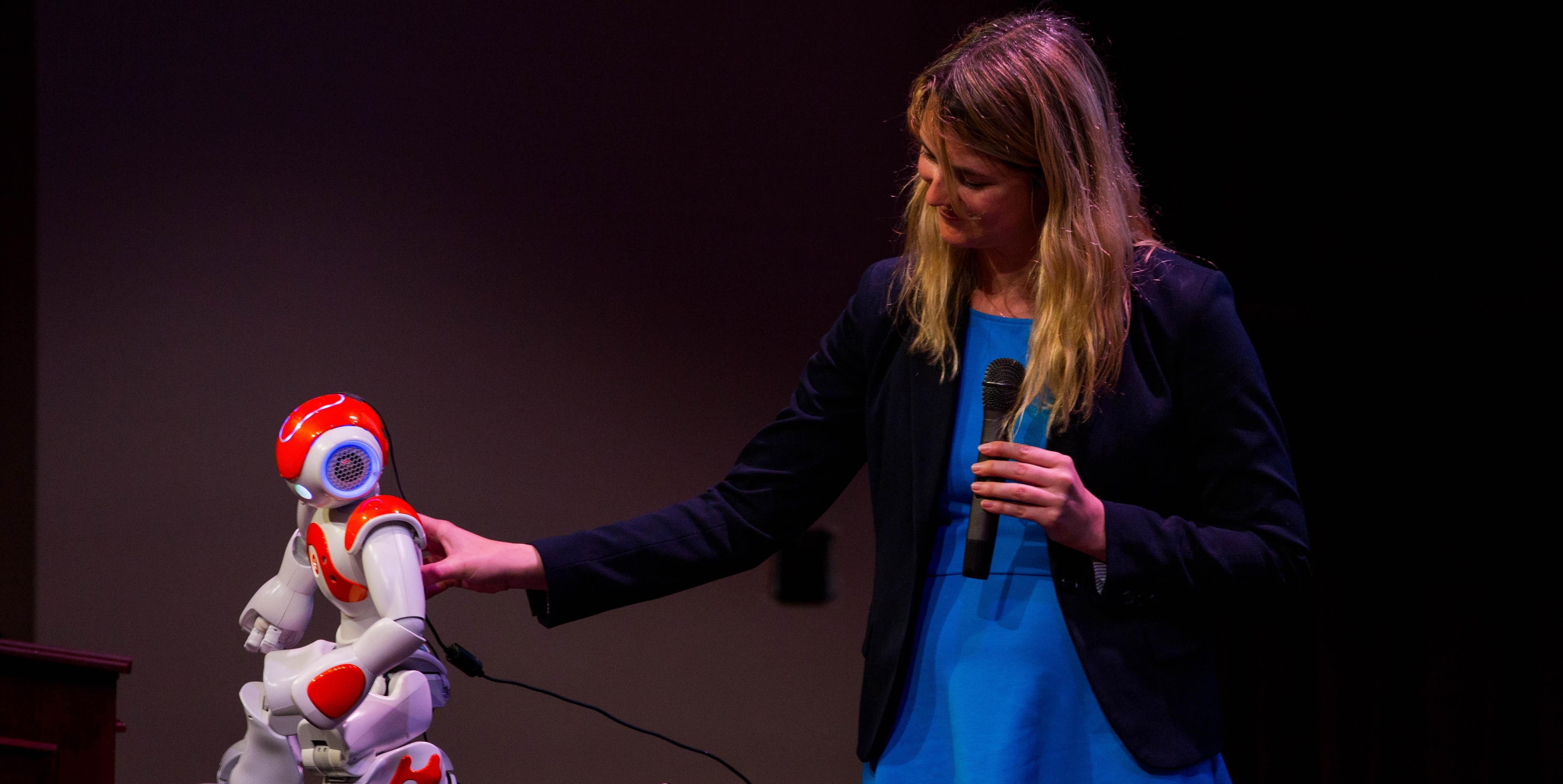 A woman holding a microphone affectionately touches a small robot.
