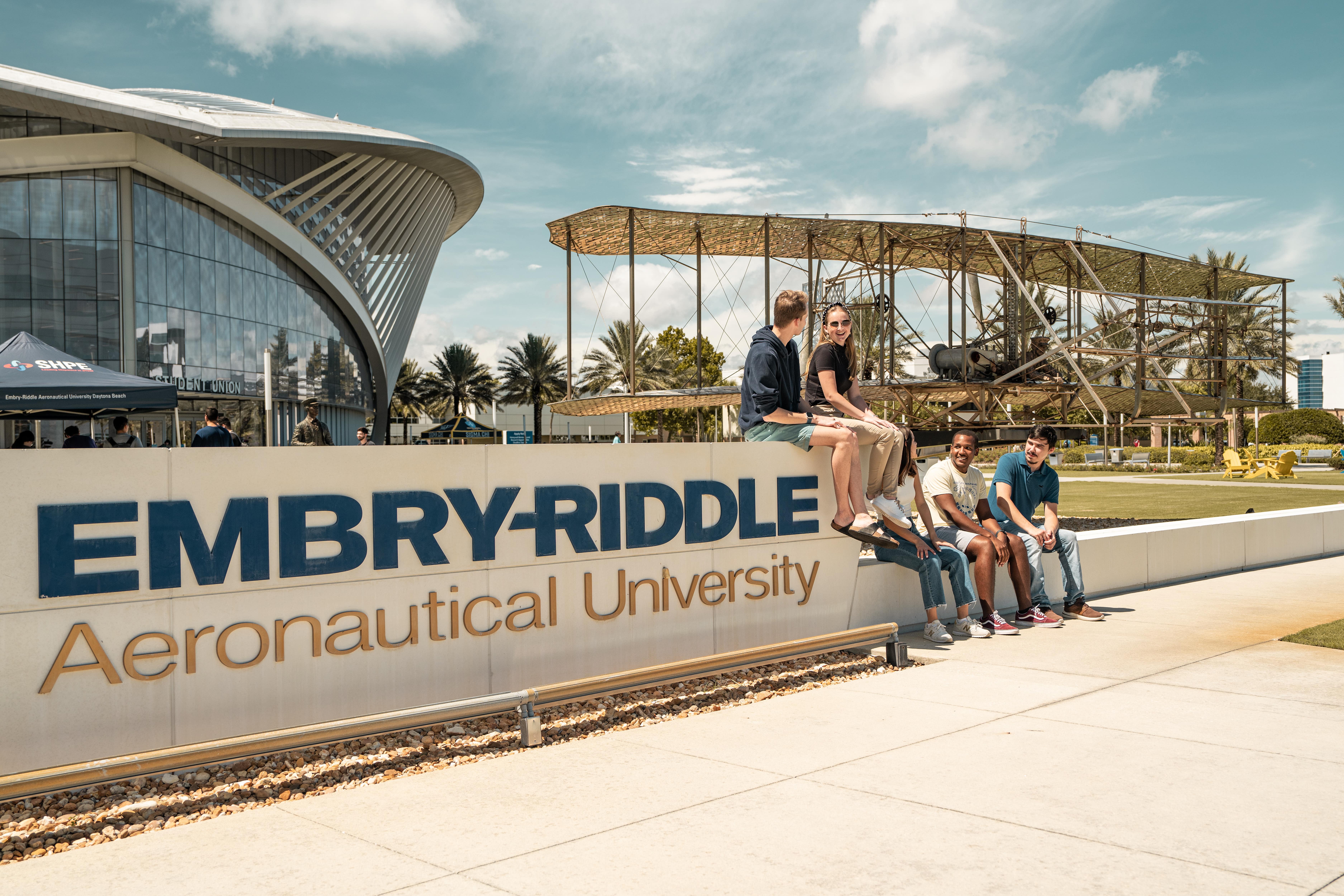 Several students sit on the Embry-Riddle monument sign at the Daytona Beach campus. A vintage biplane is seen in the background.