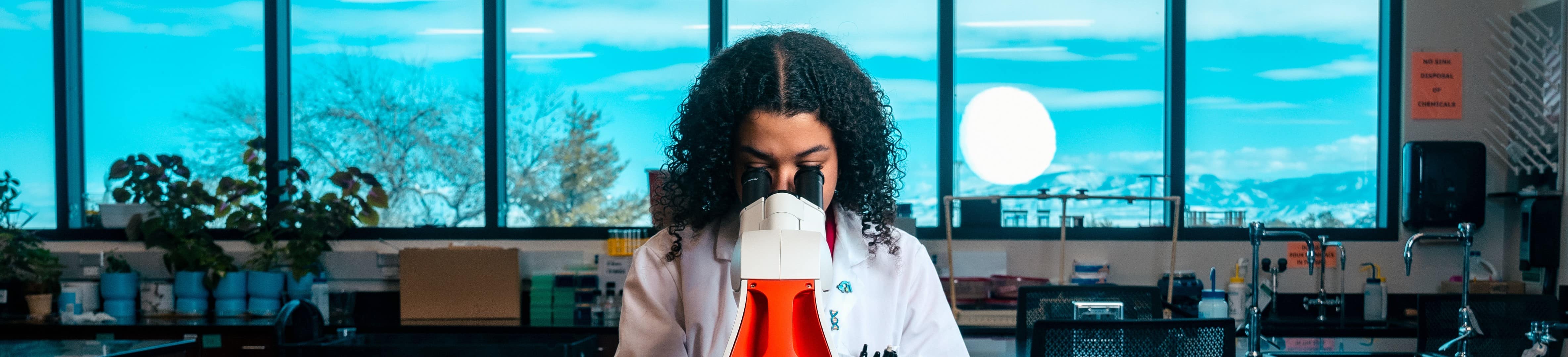 A student with dark skin tone wears a lab coat and looks into a microscope. Mountains can be seen through the wall of windows in the background.