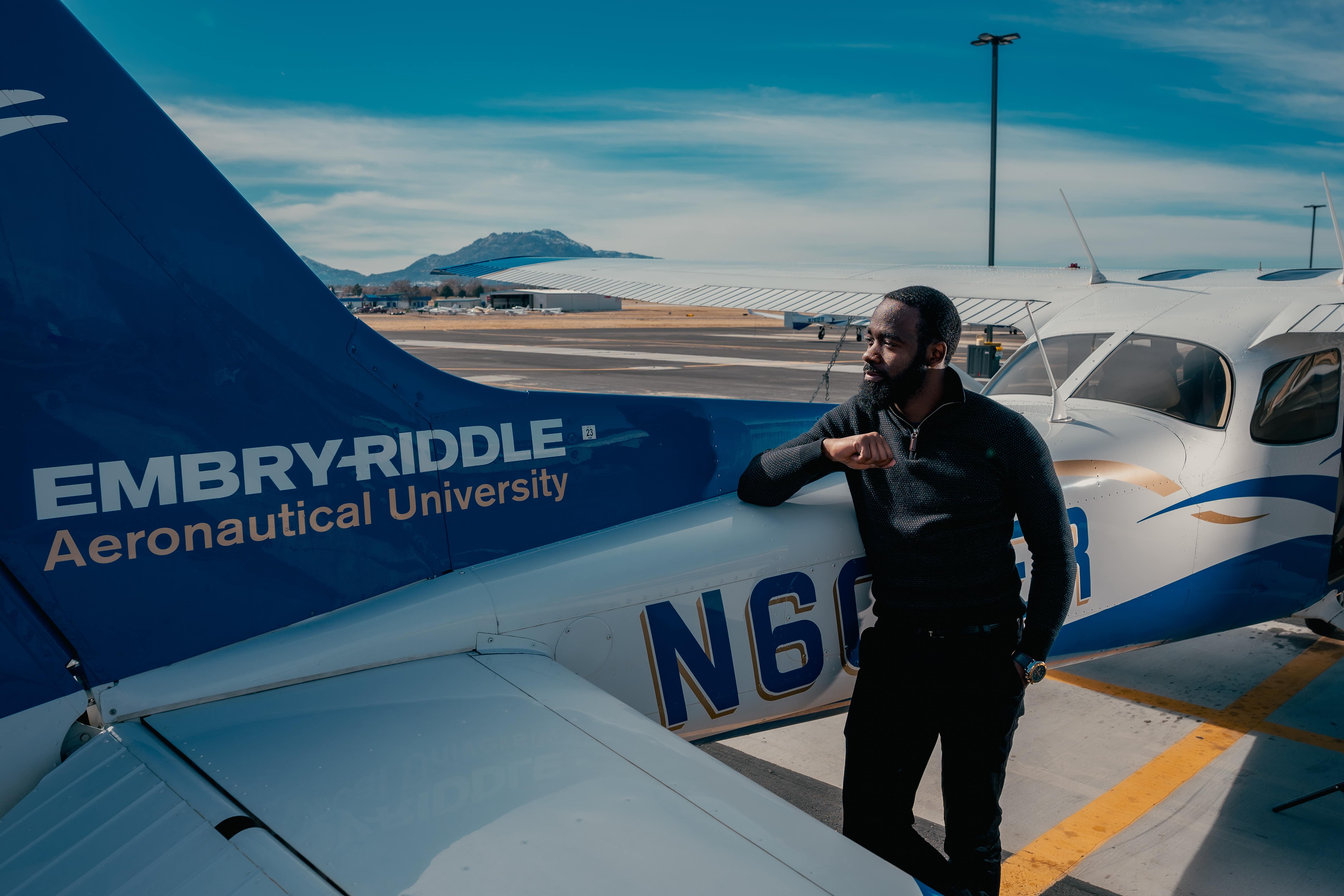 A Black student with a beard leans against a small aircraft. Prescott's Granite Mountain can be seen in the background.