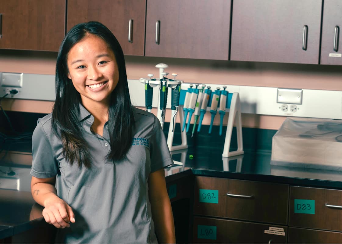 Student in a grey polo shirt smiles, standing in a lab.