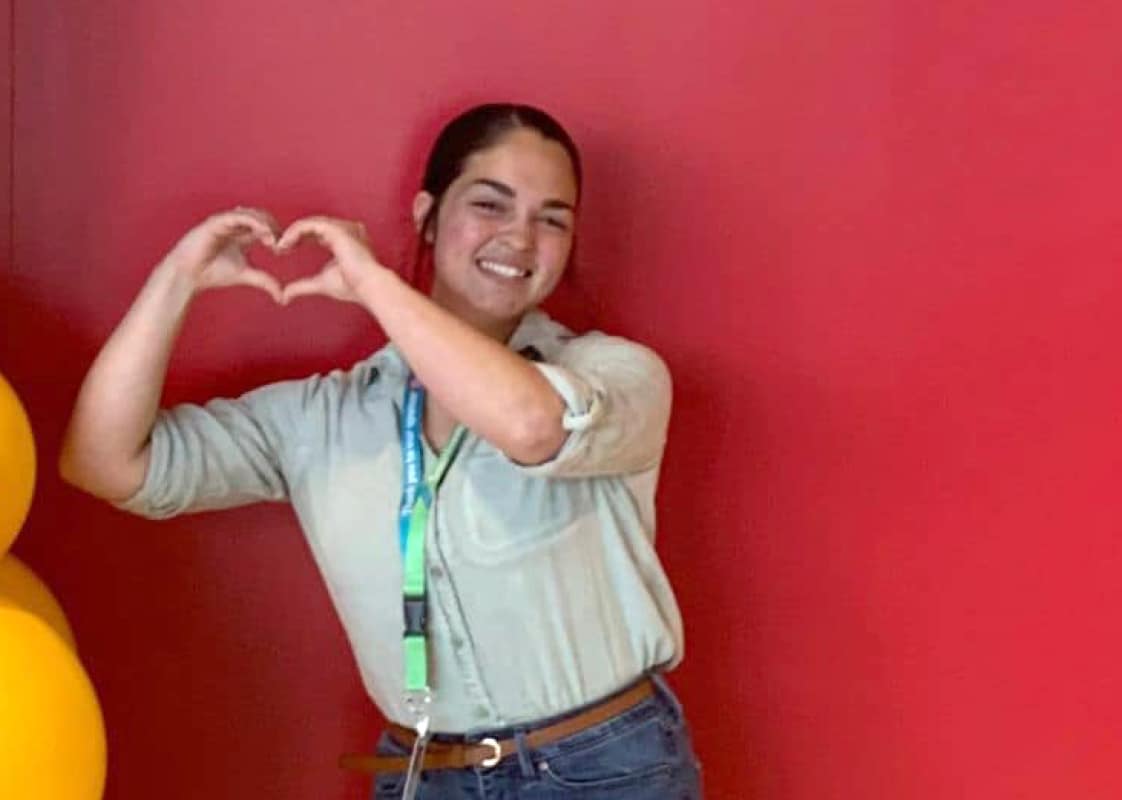 Mia Hamlin, standing in front of a red wall, holds her hands in a heart shape.
