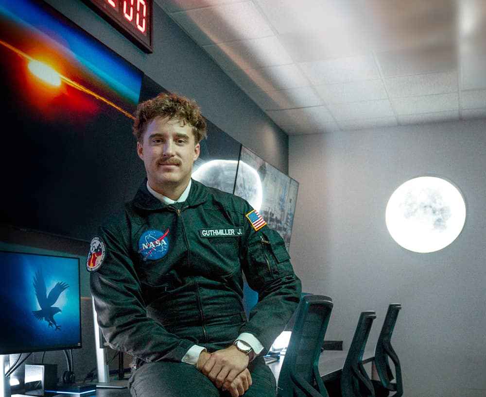A student with a mustache wearing a jumpsuit with a NASA insignia leans casually against a desk with several computer monitors in a darkened room. A glowing moon shines on the wall behind him.