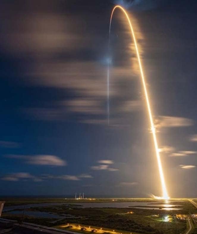 A glowing trail arcs away from the ground into the dark sky after a rocket launch.