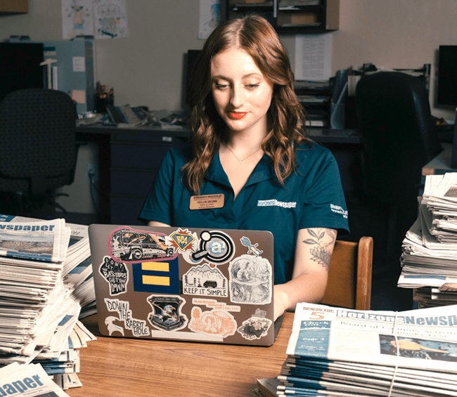 Taylor sits at her laptop, which is covered by stickers including an equals sign symbolizing equality and human rights. Stacks of newspapers cover the desk.