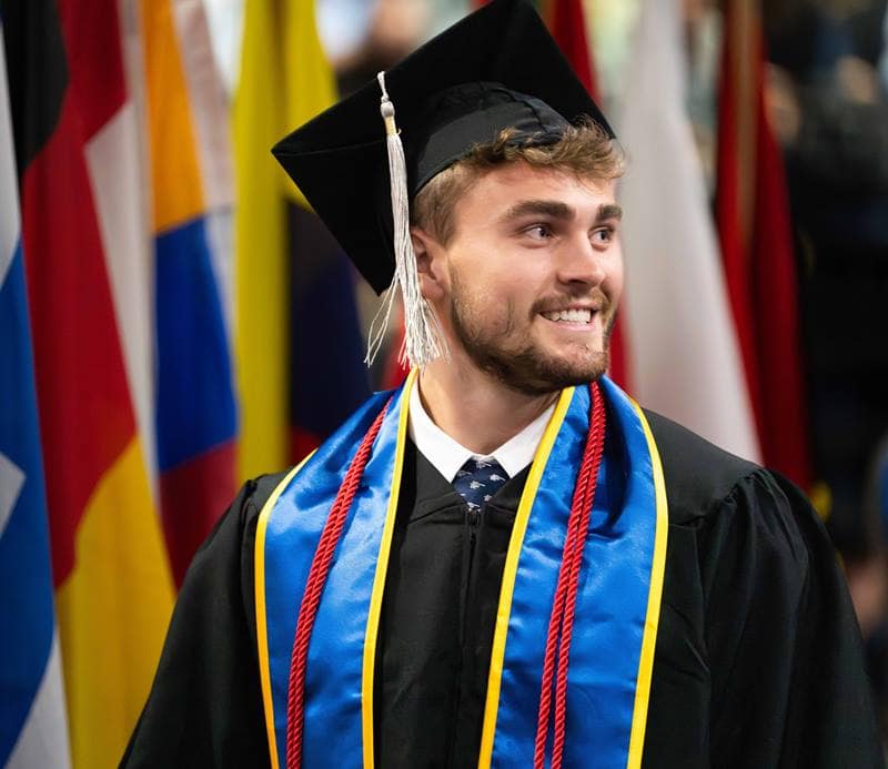 A white man with a dark blond beard wears a graduation cap and gown, a blue and yellow sash, and red cords.