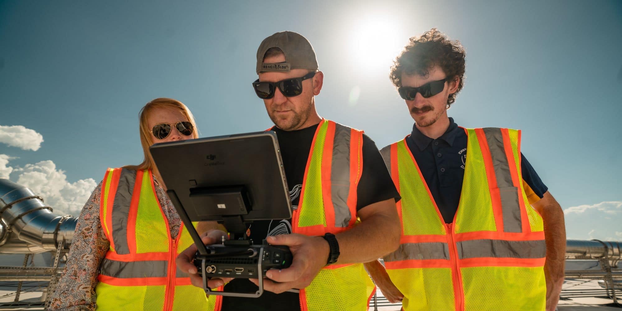 Three students in sunglasses and reflective vests look at the display of a drone control.