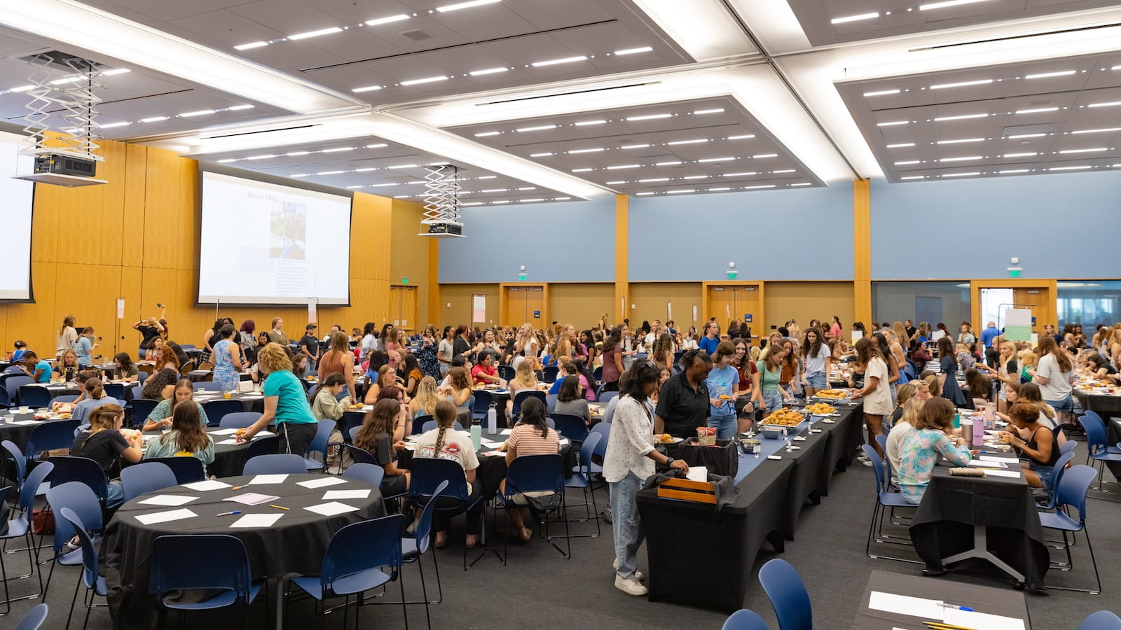 Large student networking event in a campus conference hall with round tables, buffet stations, and projection screens overhead.