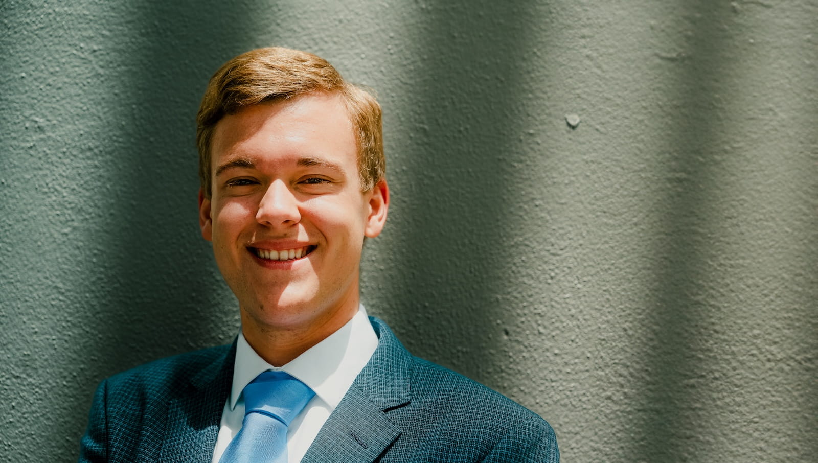 Student poses in a light blue checked blazer and light blue tie, standing against a textured exterior wall in natural light.