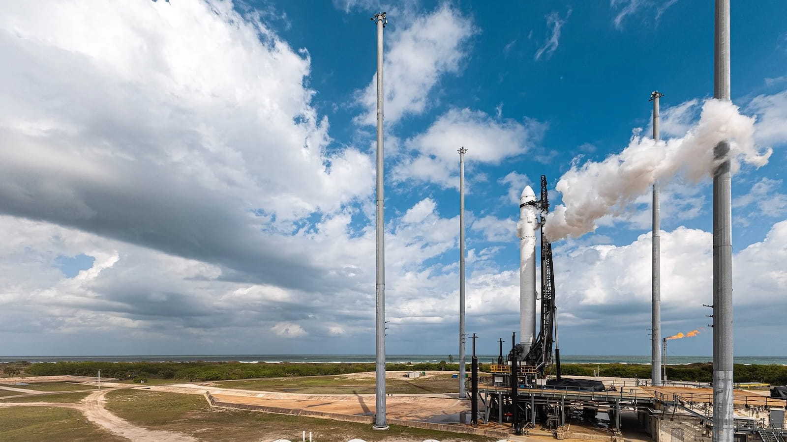 Orbital launch vehicle on a coastal launch pad venting white propellant vapor, surrounded by lightning towers and flame stack, with the ocean in the background.
