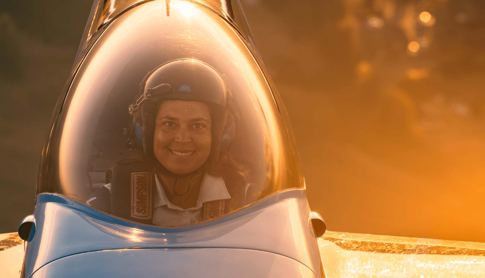 Student pilot wearing a flight helmet sits in a small aircraft cockpit with a bubble canopy, illuminated by warm sunset light.