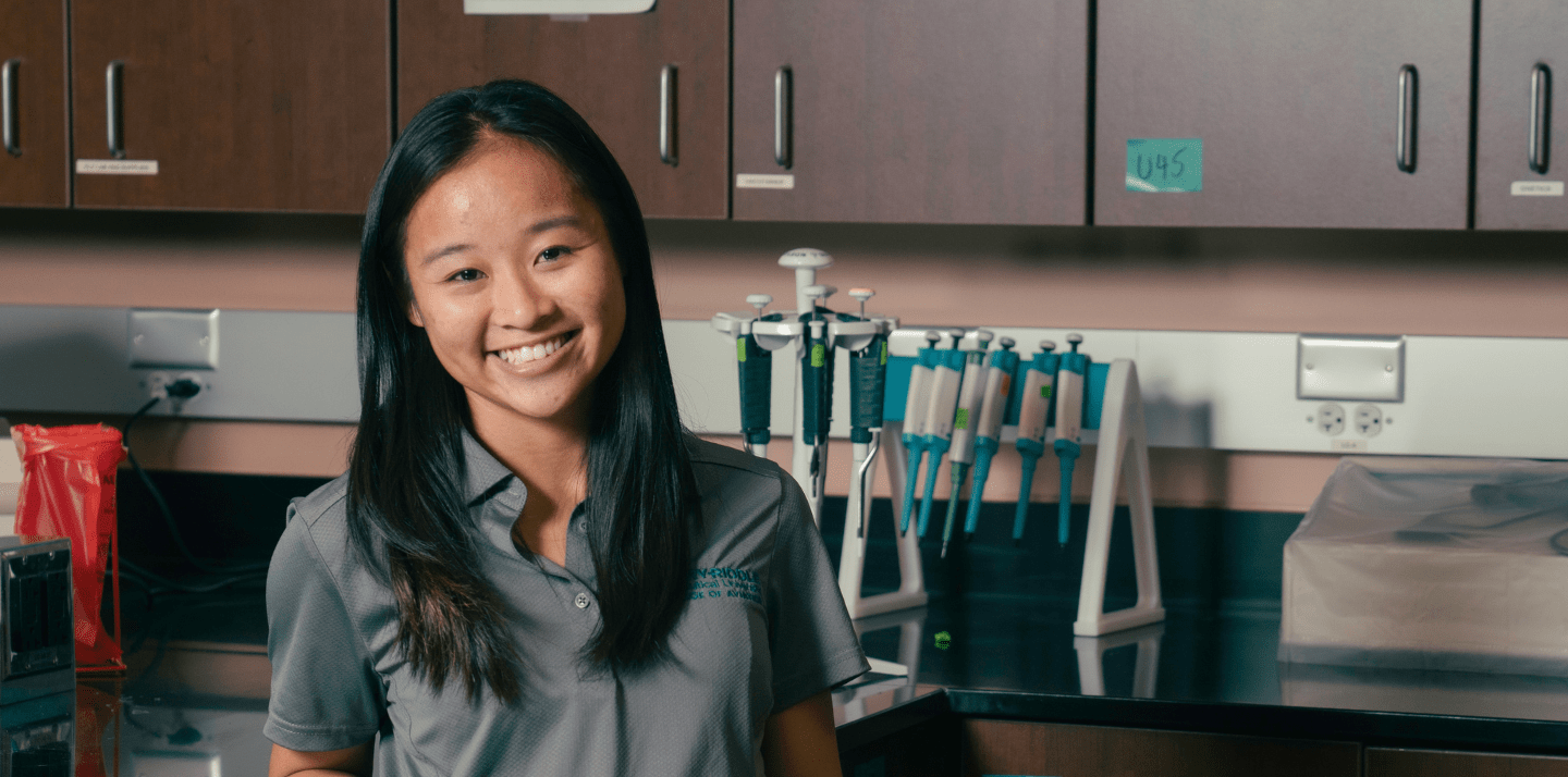 Student wearing an Embry-Riddle College of Business polo stands in a laboratory workspace with micropipettes, glassware, and lab equipment on the bench behind her.