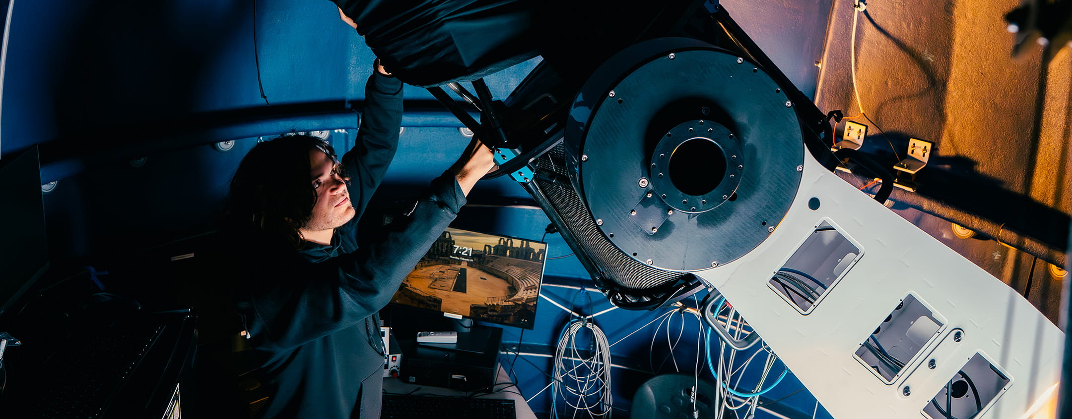 Student adjusting mounted observatory telescope with cabling and control system, inside dome with monitoring computer displaying sky or observation feed.