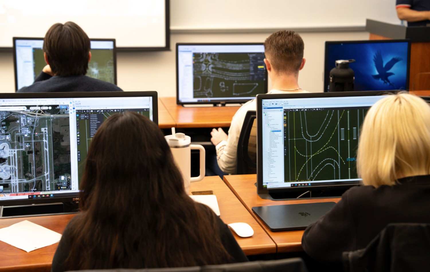 Students use GIS and mapping software on desktop monitors displaying aerial imagery and roadway design layouts in a classroom setting.