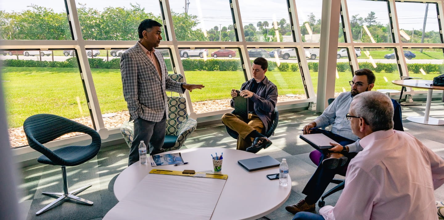 Faculty member leads a small group discussion with students seated around a table in a glass-walled campus meeting space.