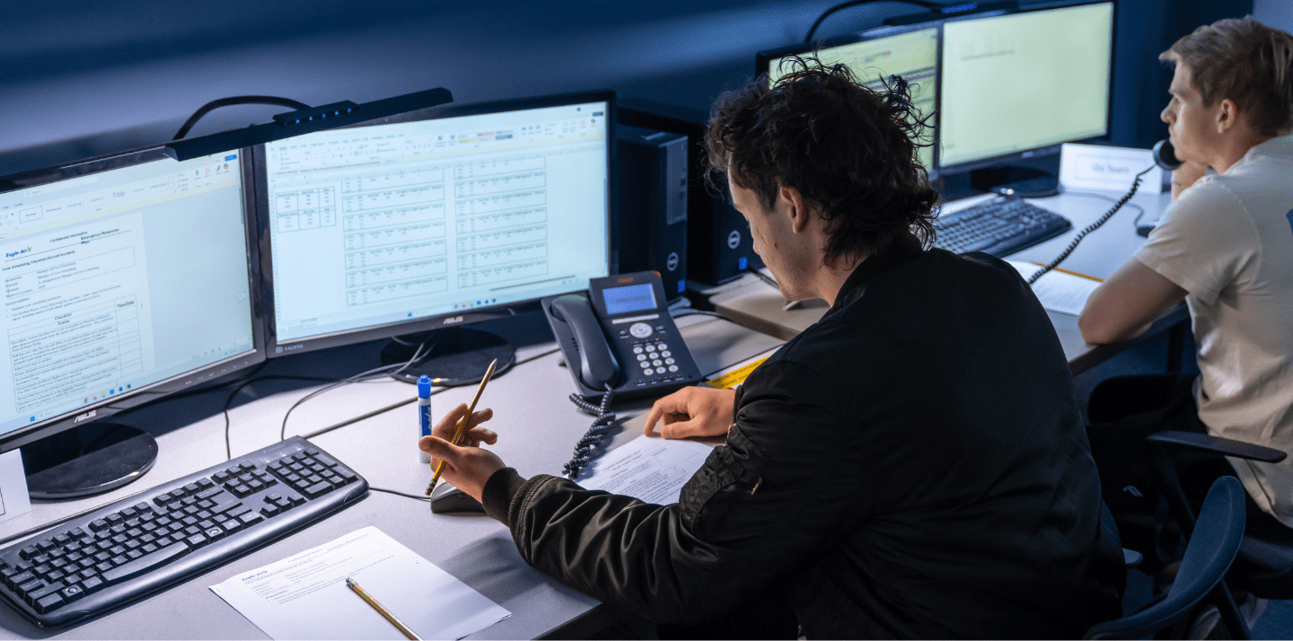 Students work at dual-monitor computer stations, reviewing spreadsheets and documents while one speaks on a desk phone in a lab setting.