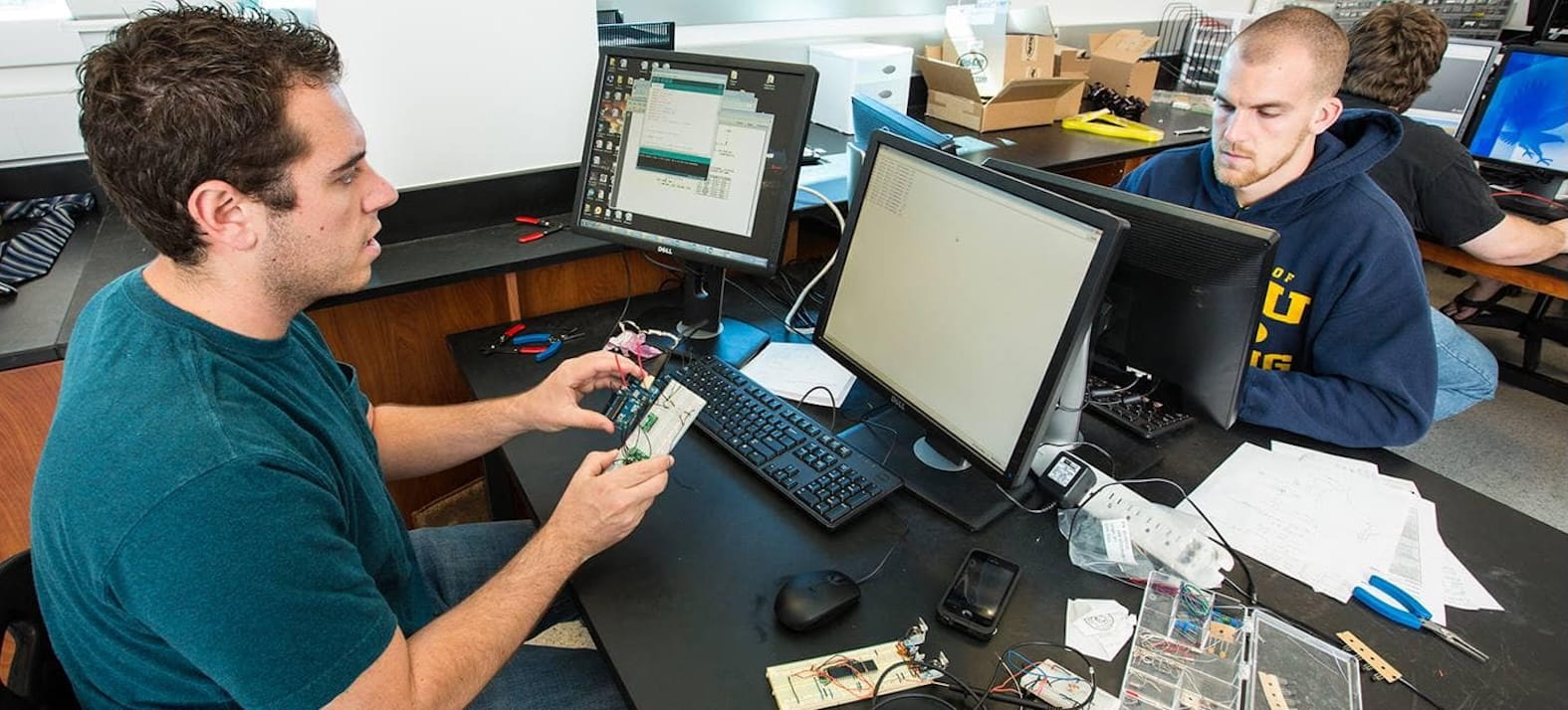 Students collaborate at a computer workstation, testing and discussing a small electronics prototype with circuit boards and wiring spread across the lab bench in a candid snapshot.