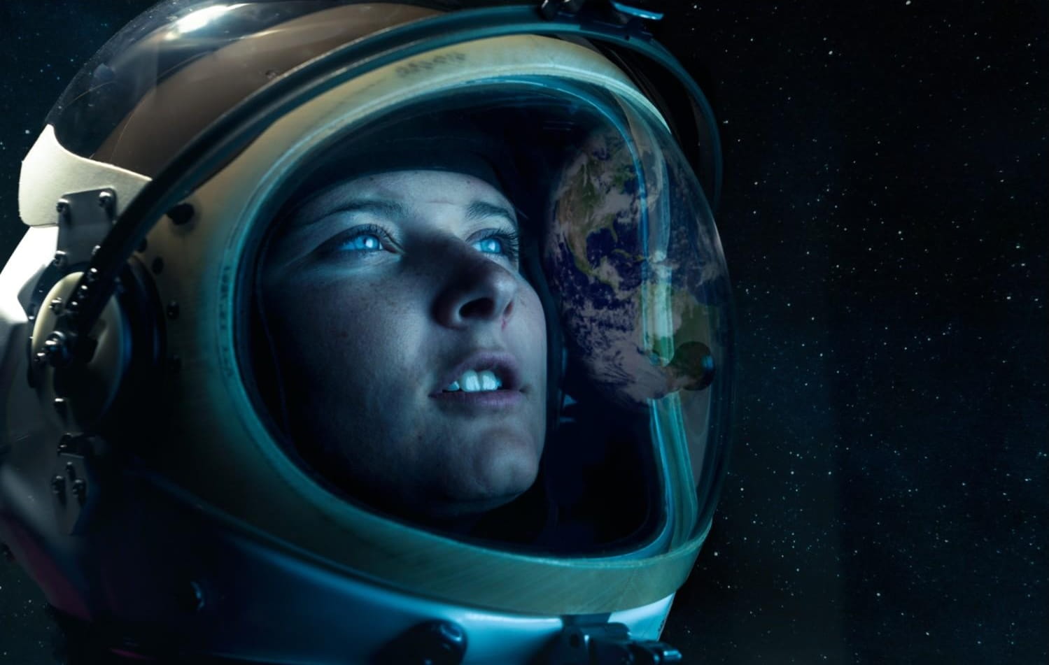 Close-up of an astronaut wearing a space helmet, gazing upward. The reflection of Earth is visible on the helmet’s visor, with a backdrop of stars in space.