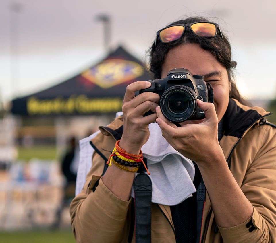 Photographer holds a Canon EOS camera up to eye, capturing an image at an outdoor campus event, with a branded tent and activity tables blurred in the background.