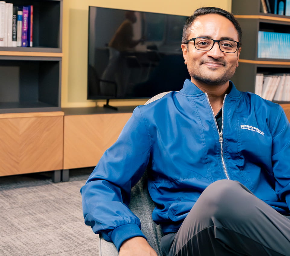 Faculty member wearing an Embry-Riddle Aeronautical University jacket sits in a lounge area with bookshelves and a television in the background.