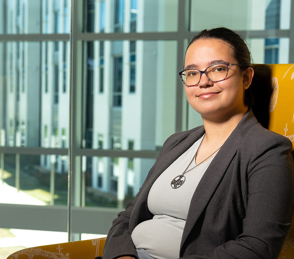 Student wearing glasses and a blazer sits in a lounge chair in front of large windows overlooking a campus building.