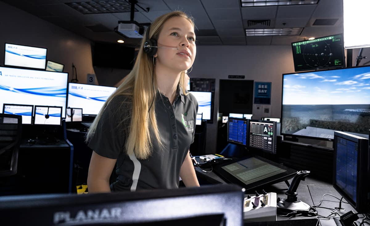 Woman wearing a headset operates controls in an aviation simulation lab surrounded by multiple monitors.