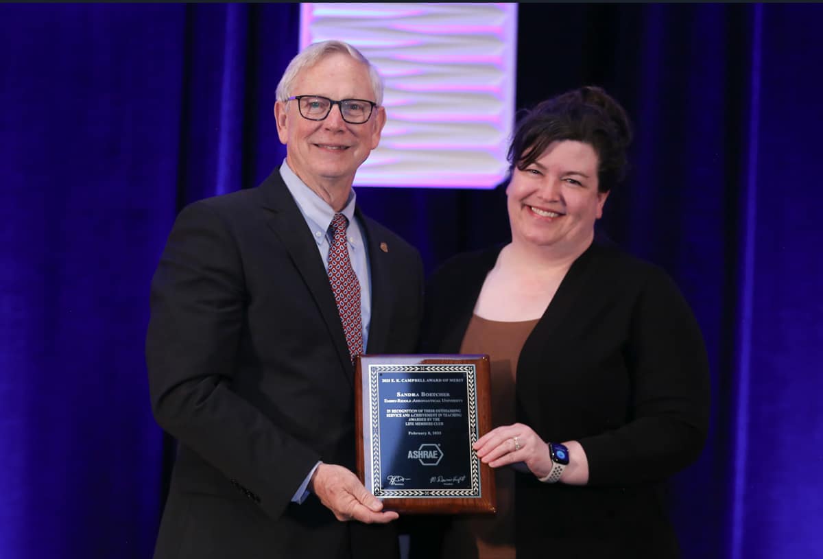 Professor Boetcher, in a dark blazer and brown top, is presented a plaque on a stage in front of a deep blue curtain.