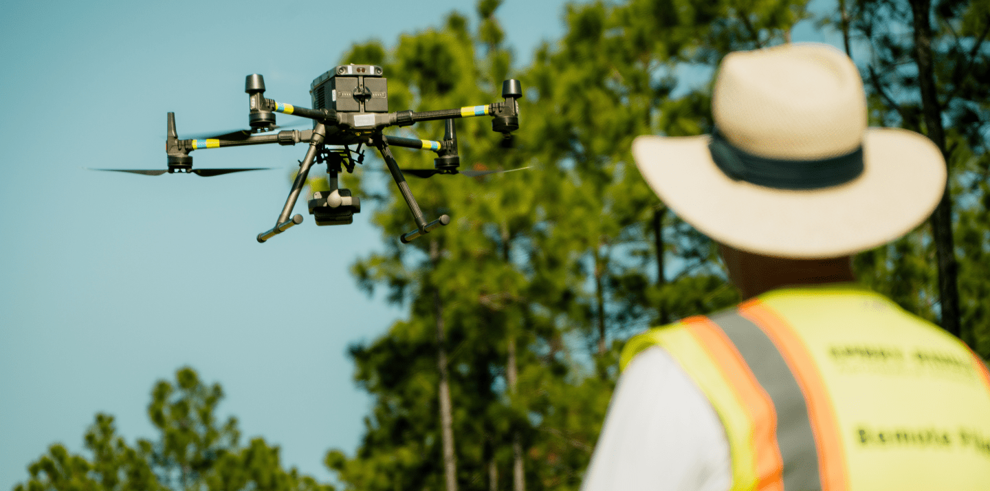 Drone hovering in front of a person wearing a safety vest and wide-brim hat in a wooded outdoor area.