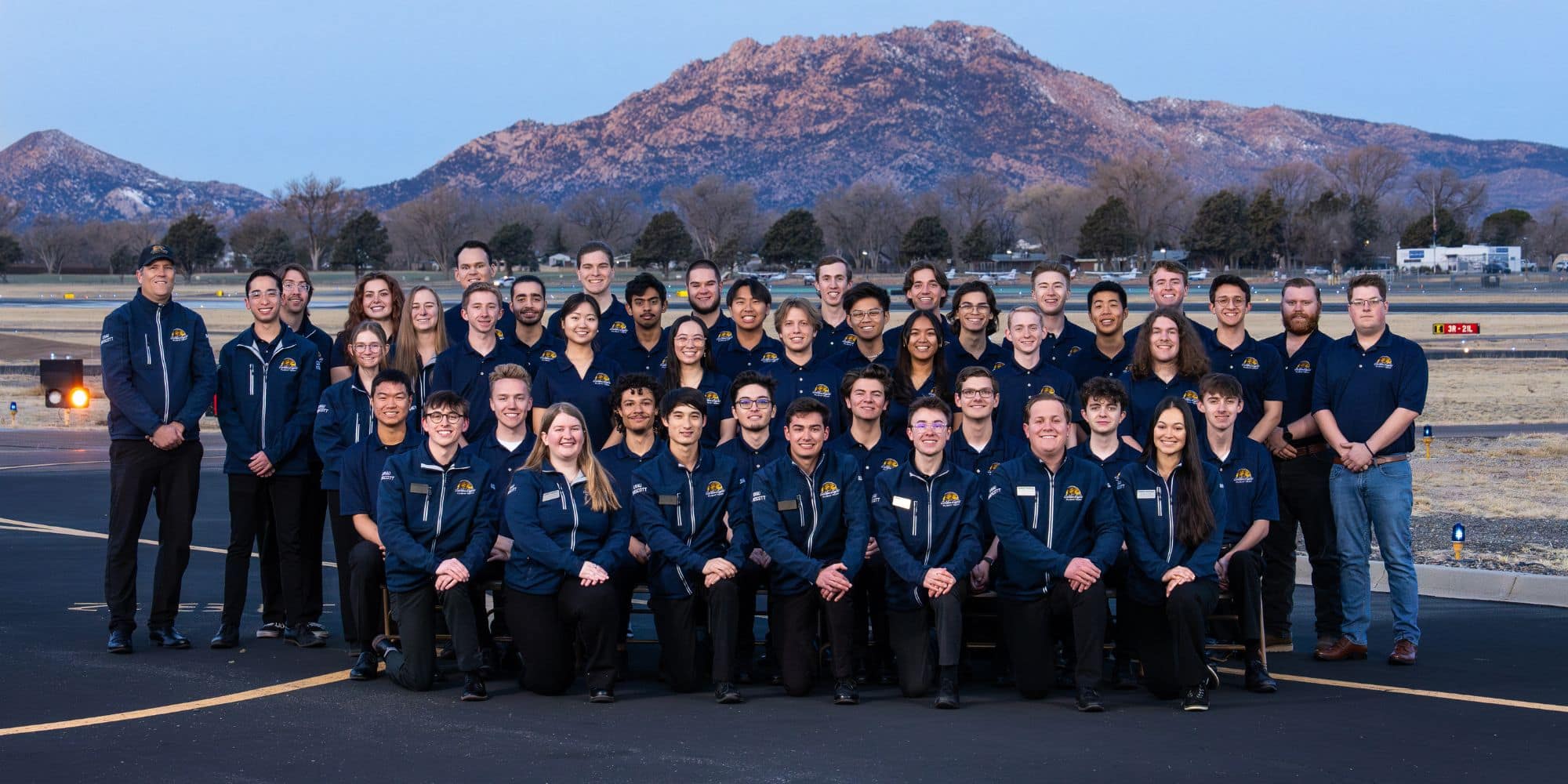 A large group of students and staff in matching navy jackets and polos with the Golden Eagles logo, pose on an airfield with mountains in the background.