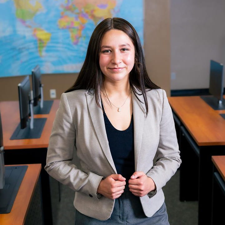 Student, Faith Vasquez stands in the Global Security Operations Center with maps on the wall behind her and computers around on desks.