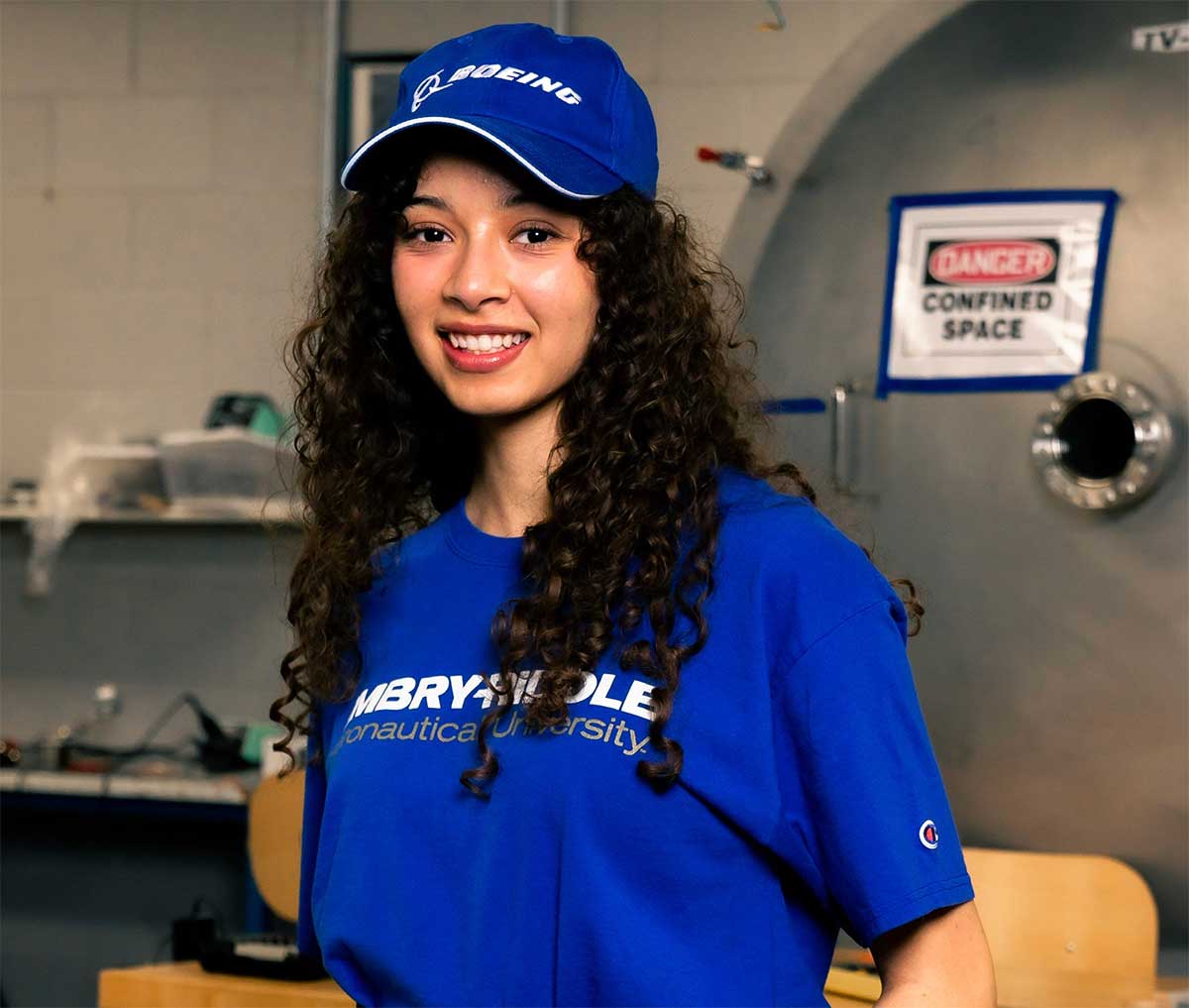 Student in Embry-Riddle T-shirt and Boeing cap smiles in a lab with equipment and a 'confined space' warning sign in the background.
