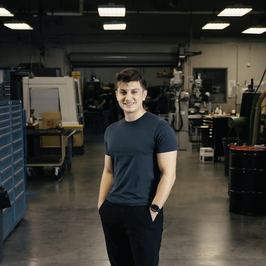A student stands smiling in a machine shop surrounded by industrial equipment.