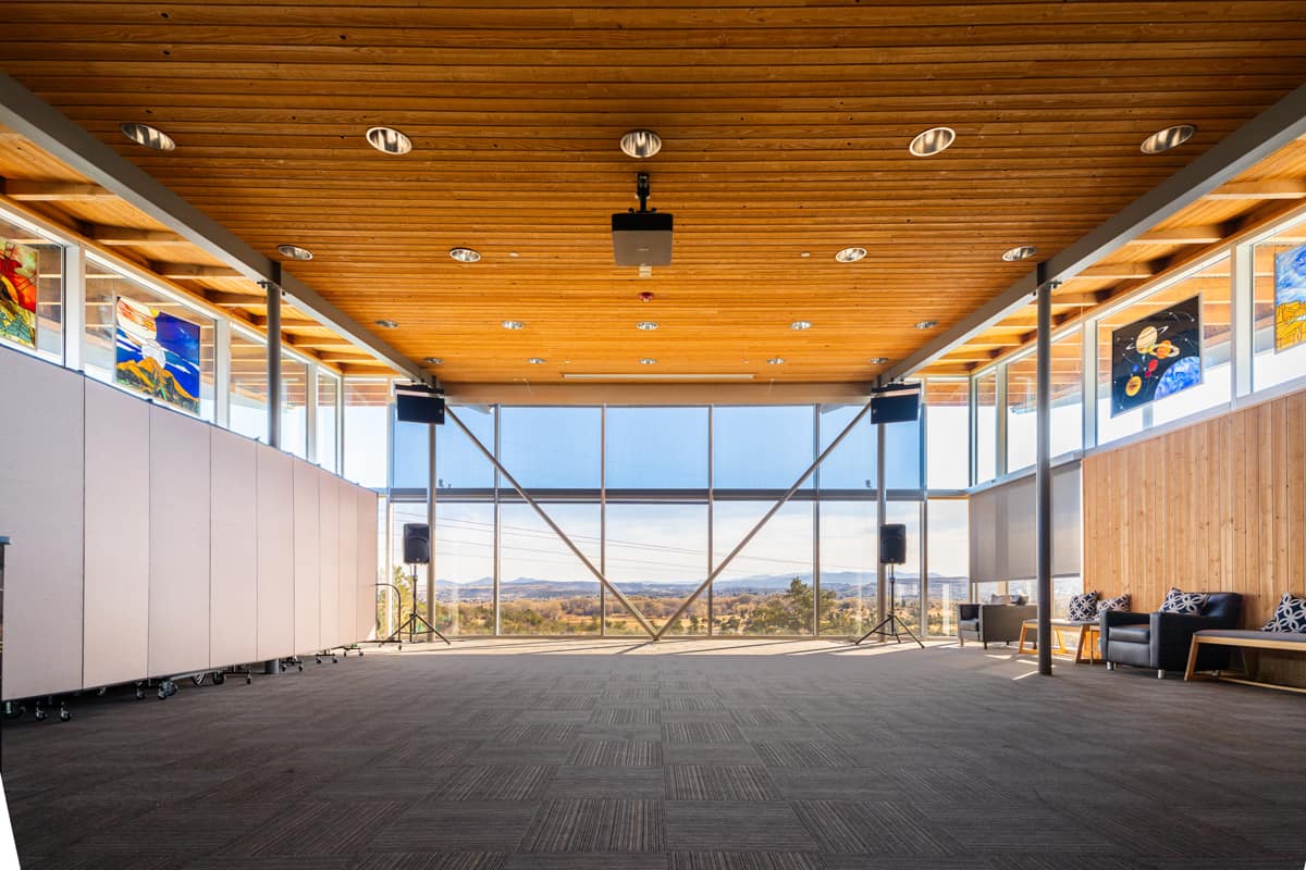 A wide, front-facing view of an empty gymnasium interior with a high wooden ceiling, large floor-to-ceiling windows.