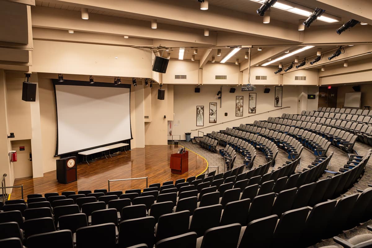 An empty auditorium with tiered seating arranged in a curved, arch-like formation and a large screen positioned on the stage.