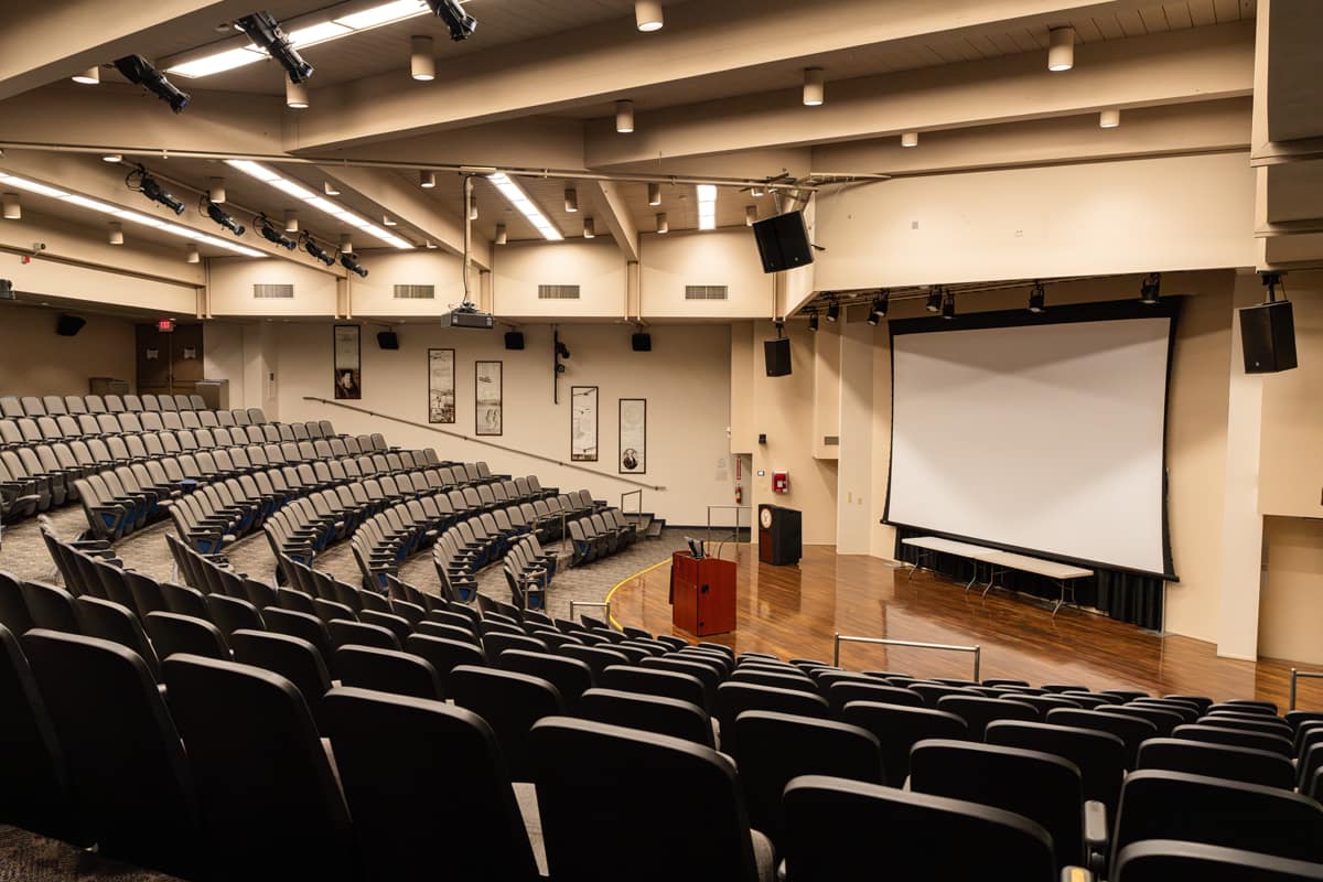 An empty auditorium with tiered seating arranged in a curved, arch-like formation and a large screen positioned on the stage.