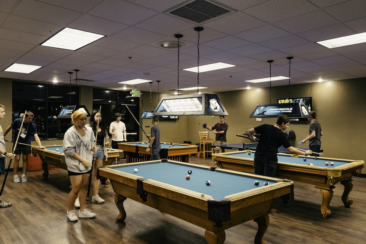 Students playing pool and table tennis in campus recreation lounge with multiple billiards tables and overhead lighting fixtures.