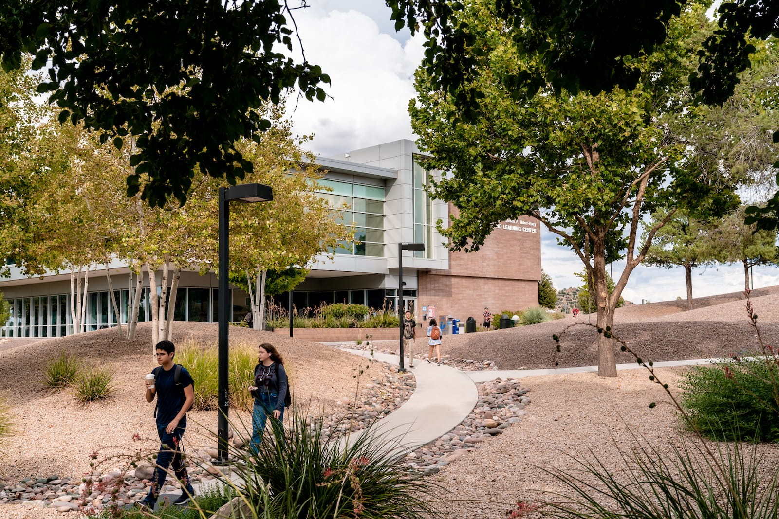 Modern campus learning center with glass facade and brick exterior, students walking along landscaped path with desert plants and trees.